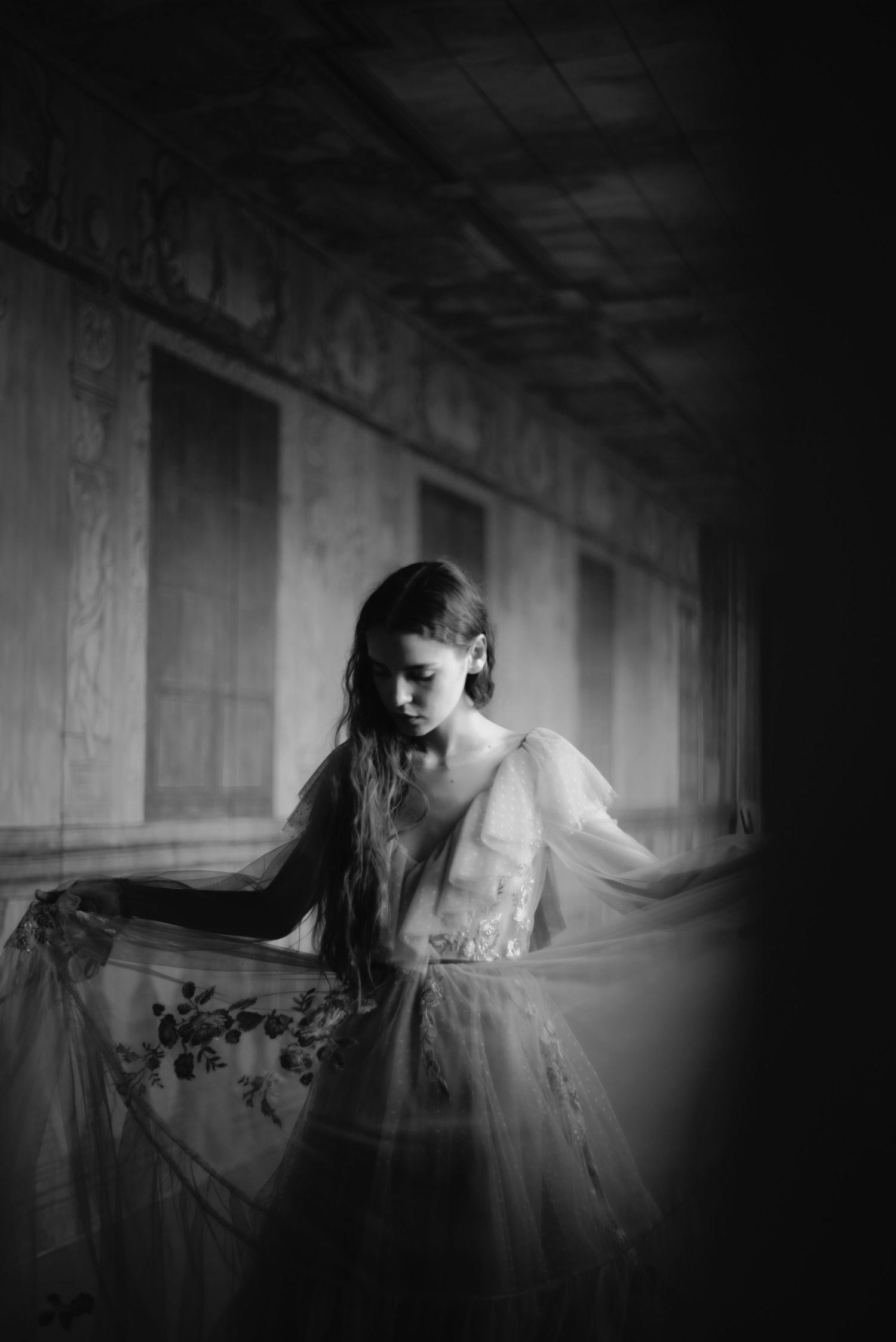 A figure in a ethereal white dress stands in a dark hallway creating a haunting and dreamy black and white image.