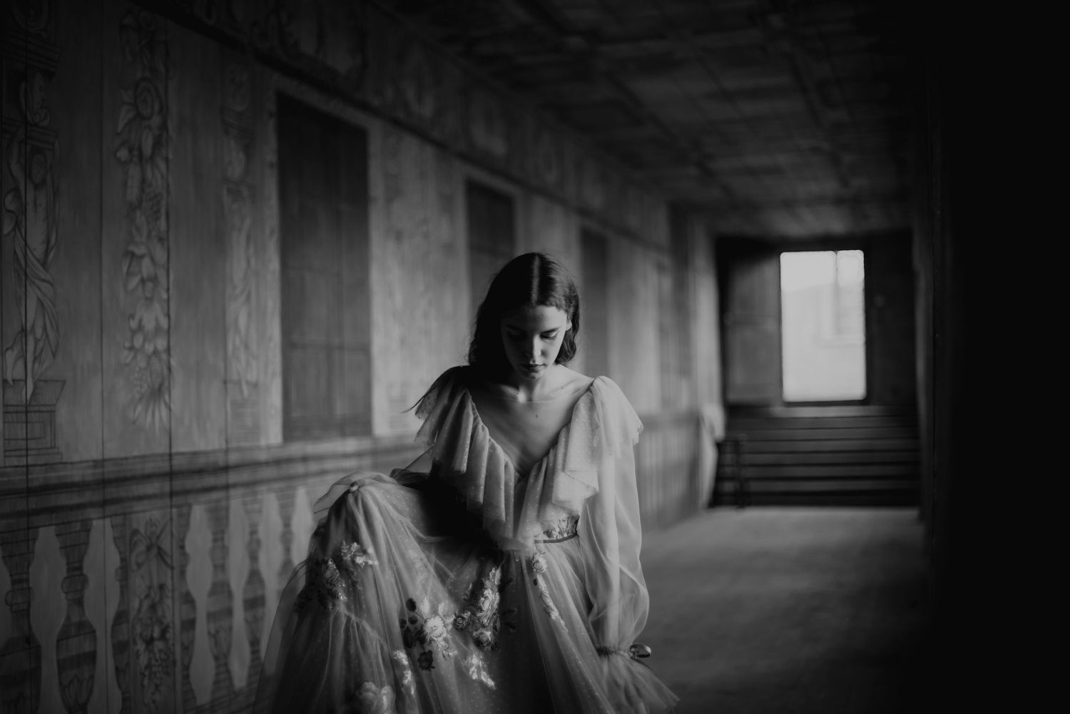 A black and white photo shows a figure in a vintage dress sitting along a hallway with ornate wood paneling.