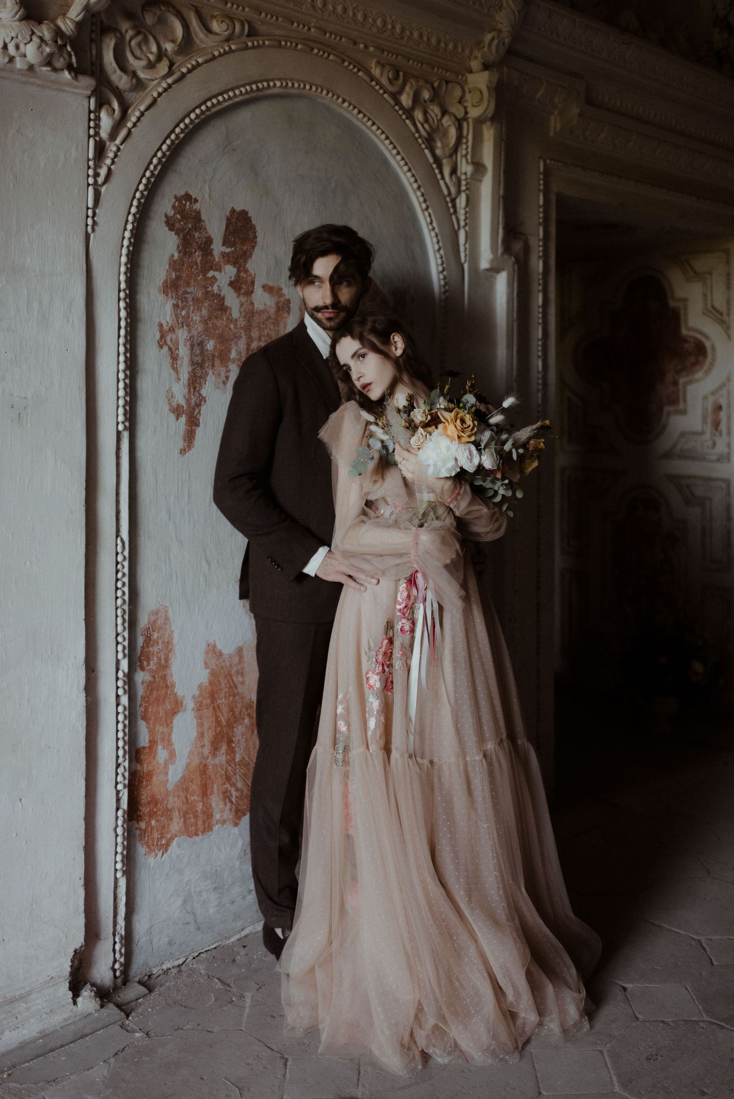 A romantic couple in vintage wedding attire pose in a moody atmospheric setting with distressed walls and ornate framing.
