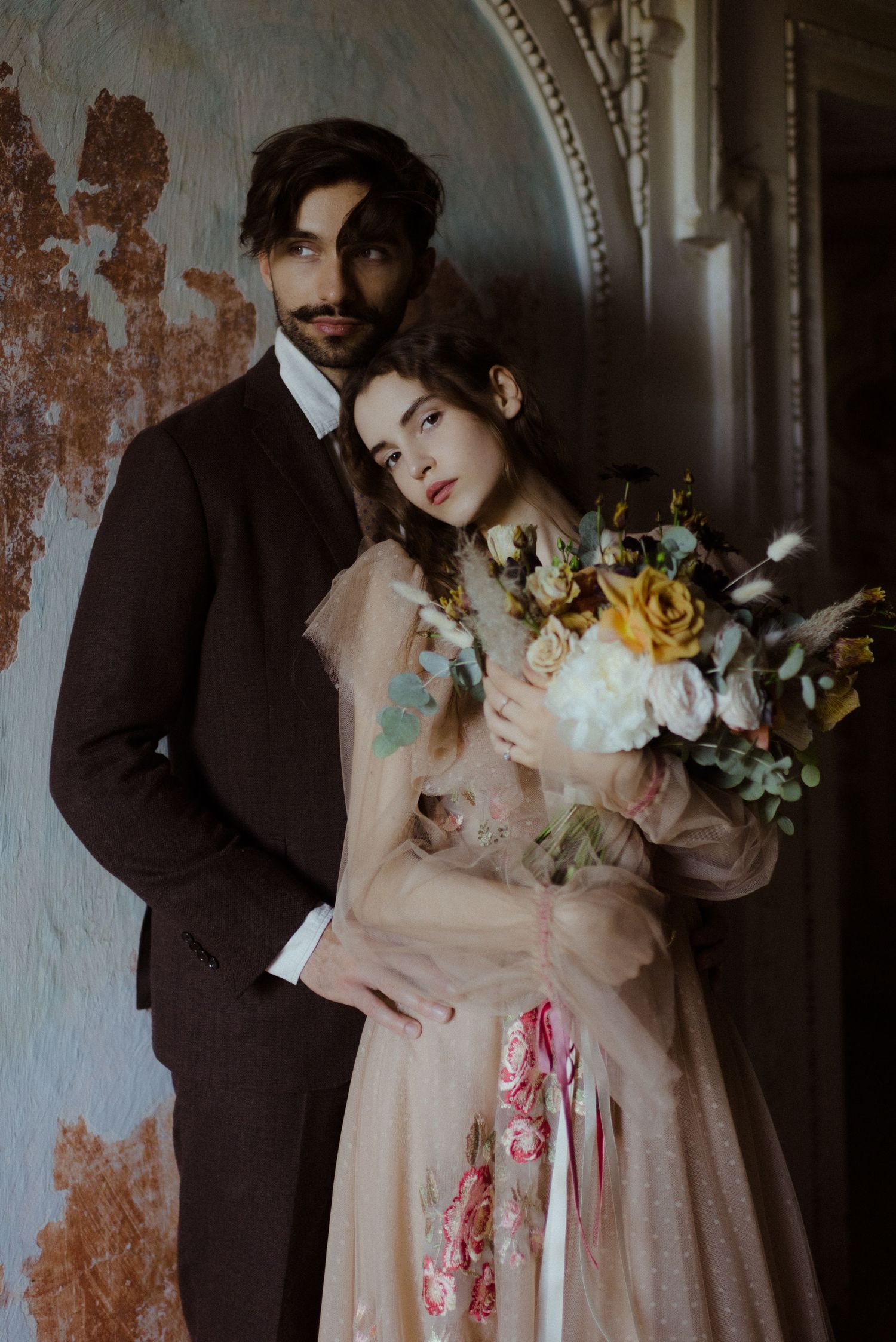 Romantic vintage-style wedding photo with couple in formal attire holding yellow and white floral bouquet against weathered wall.