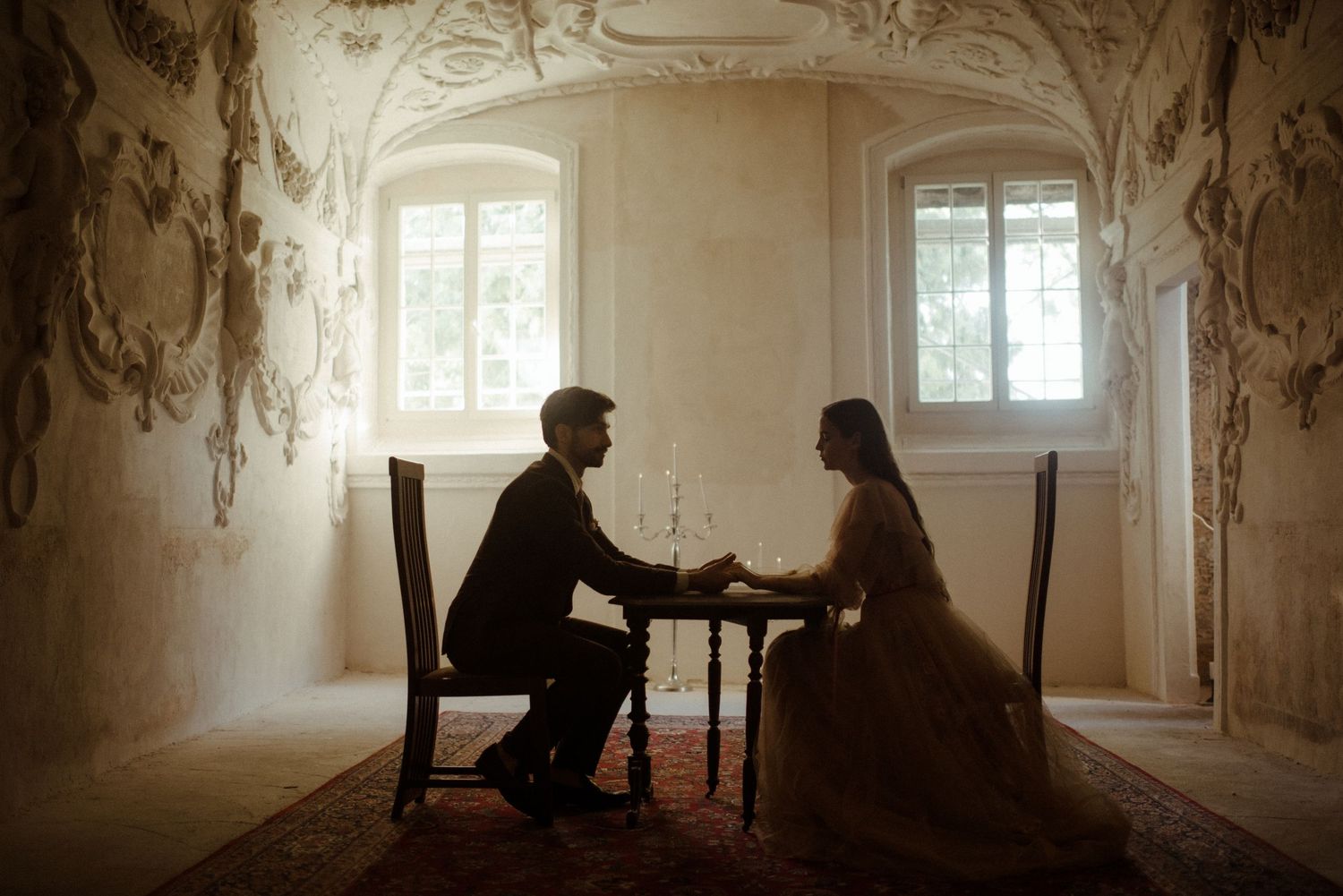 Two silhouettes sitting across from each other at a table in an elegant room with bright windows and ornate walls.