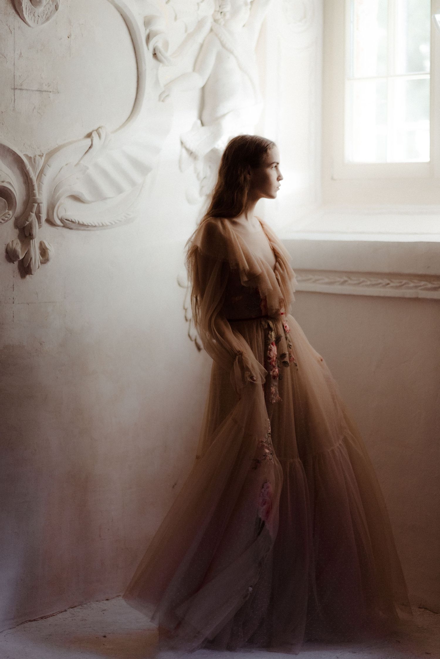 A woman in a flowing brown tulle gown stands in profile by an ornate white wall near a window.