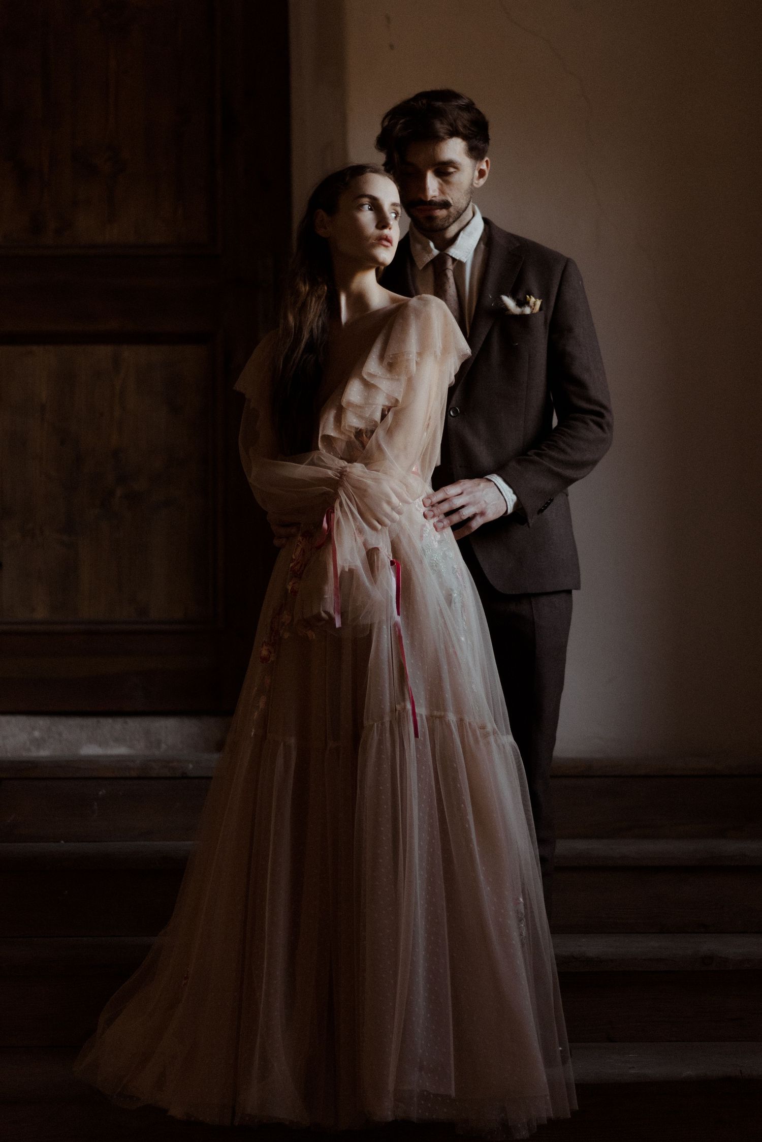A moody vintage-style wedding portrait shows a couple in formal attire with a bride in a flowing tulle gown.