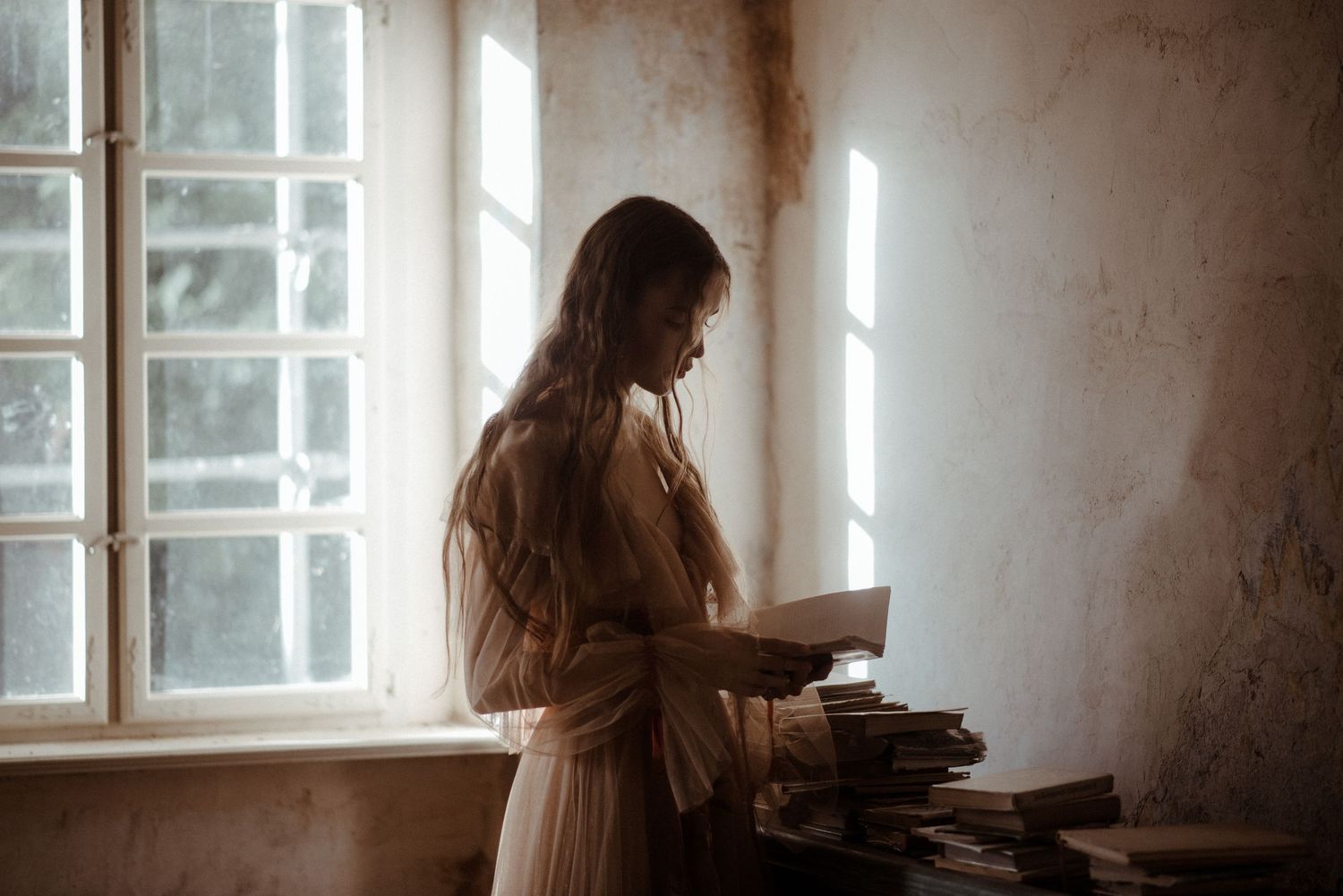A silhouetted figure in a vintage dress stands by a window reading from a stack of old books.