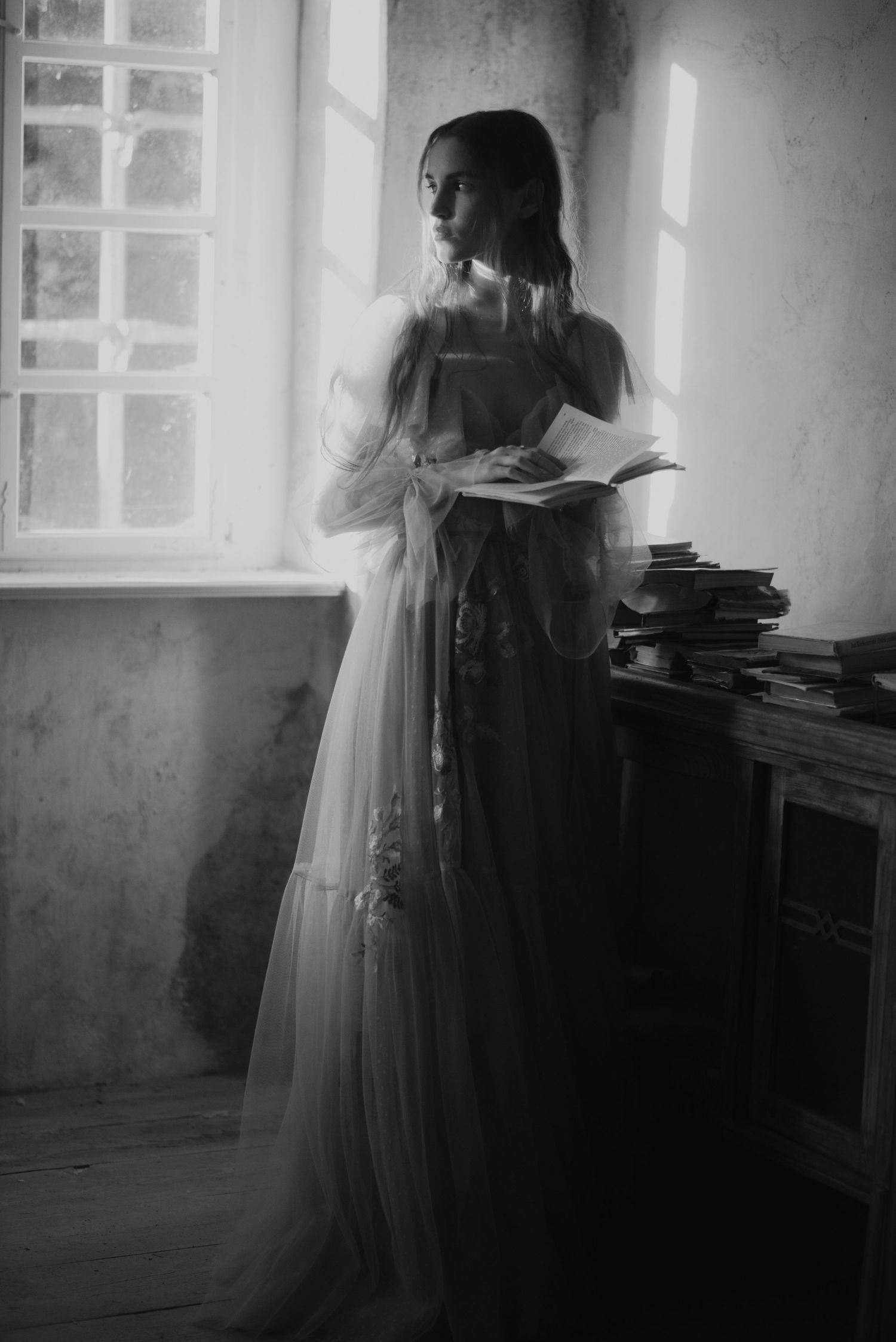 A dreamy black and white photograph of someone in a long vintage dress reading by a window with stacked books nearby.