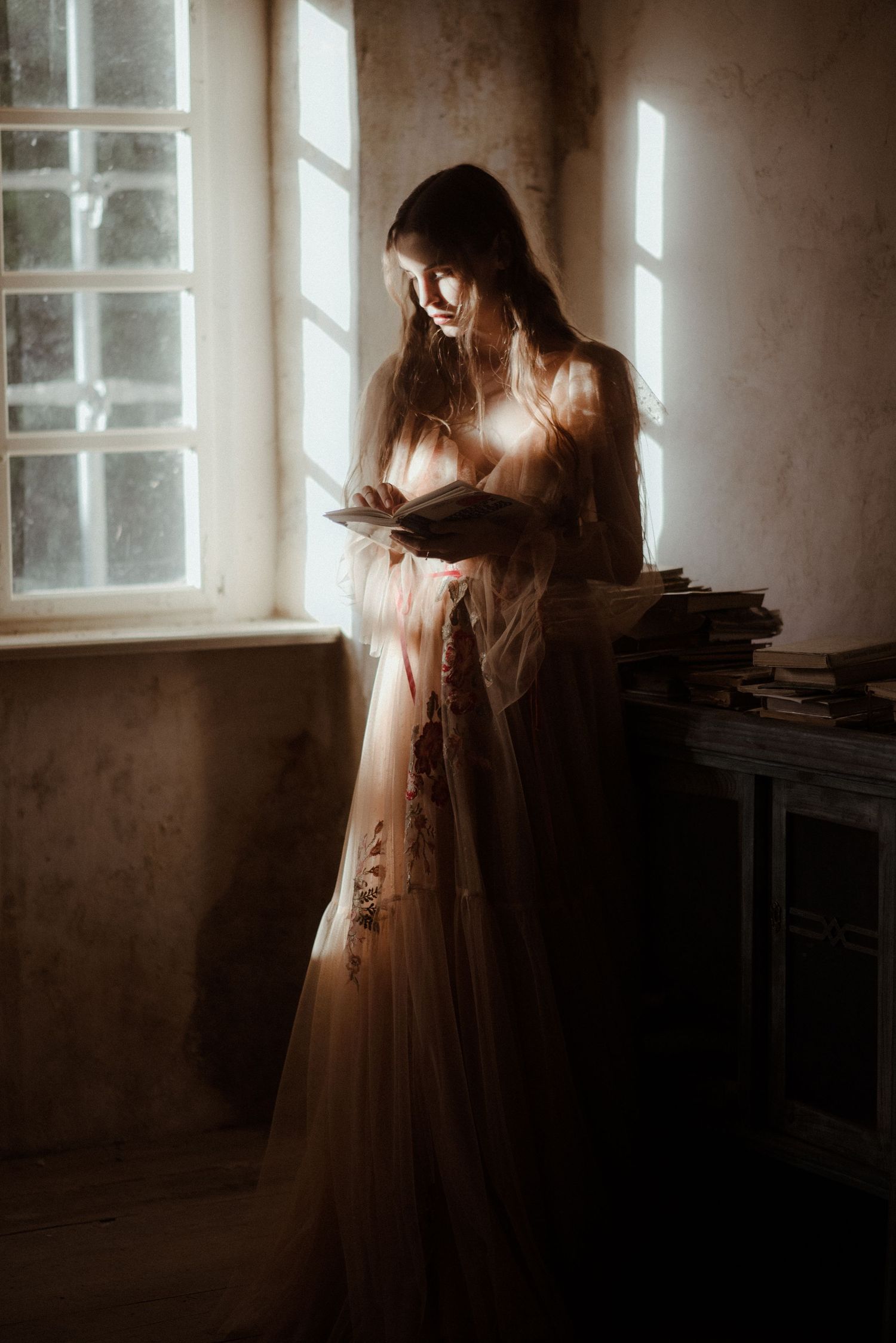 A silhouetted figure stands in ethereal light by a vintage window, wearing a flowing dress while reading in a moody atmosphere.