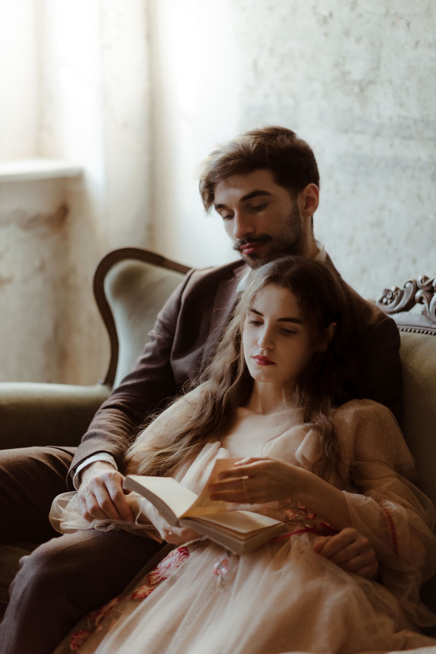 A couple relaxes together on an antique sofa while reading a book in soft natural window light.