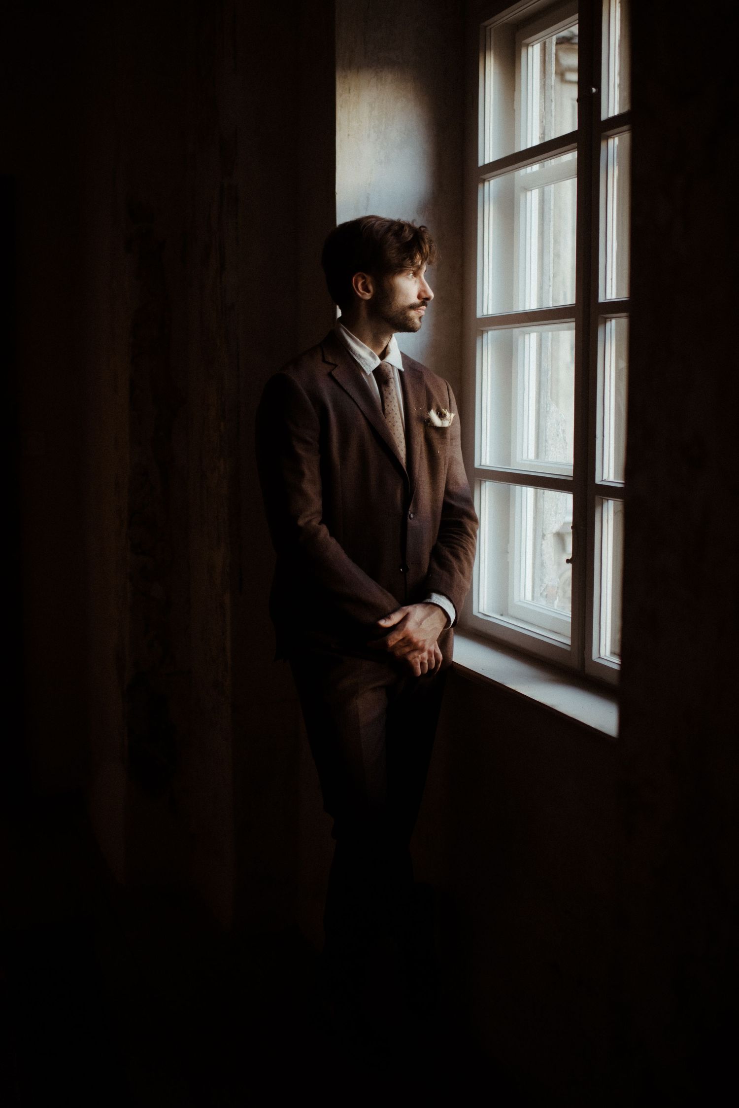A person in a brown suit stands contemplatively by a window in a dark moody room with natural light streaming in.