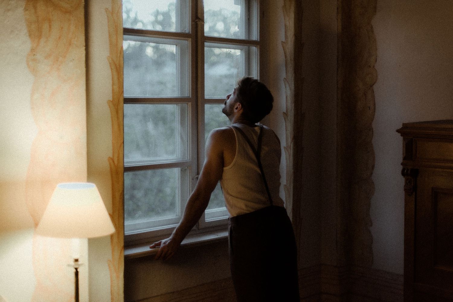 Silhouette stands by window in dimly lit room with warm lamp glow in evening light.