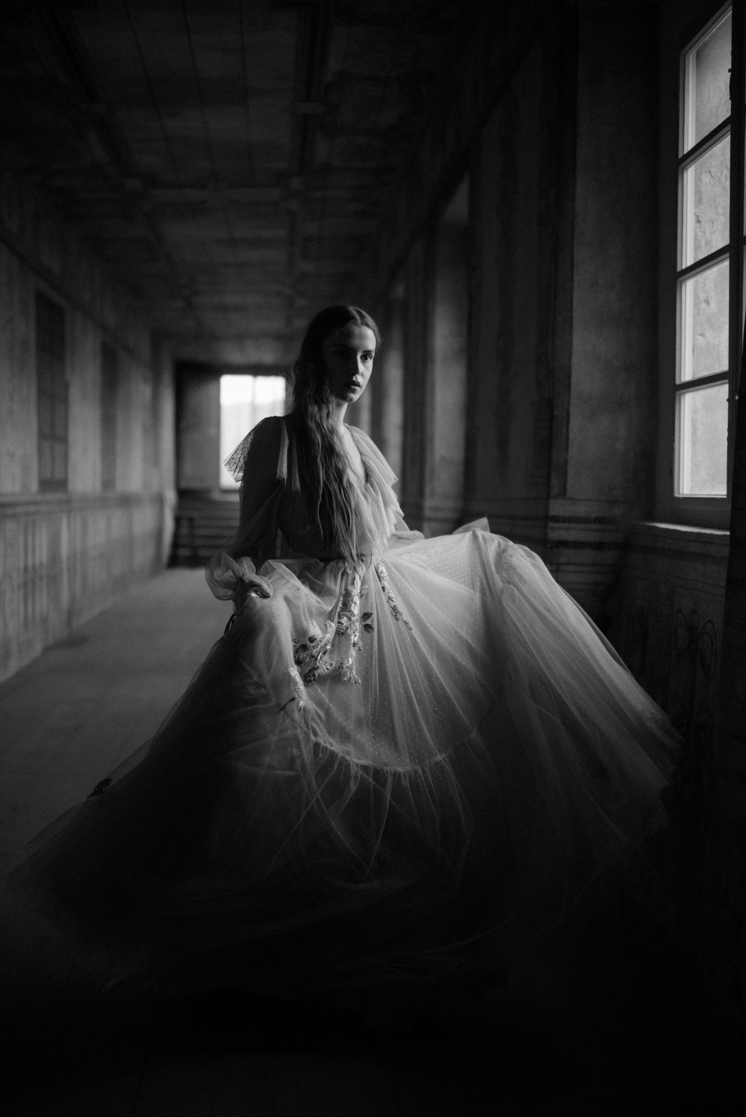 A figure in a flowy white dress twirls dramatically in a shadowy corridor lit by window light.