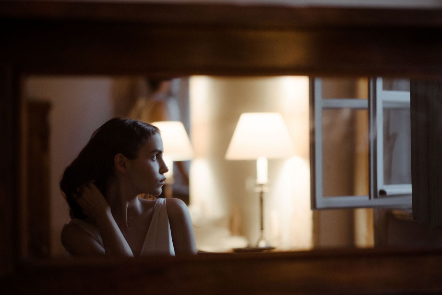 A silhouetted figure sits pensively in a dimly lit room with a warm glowing table lamp in the background.