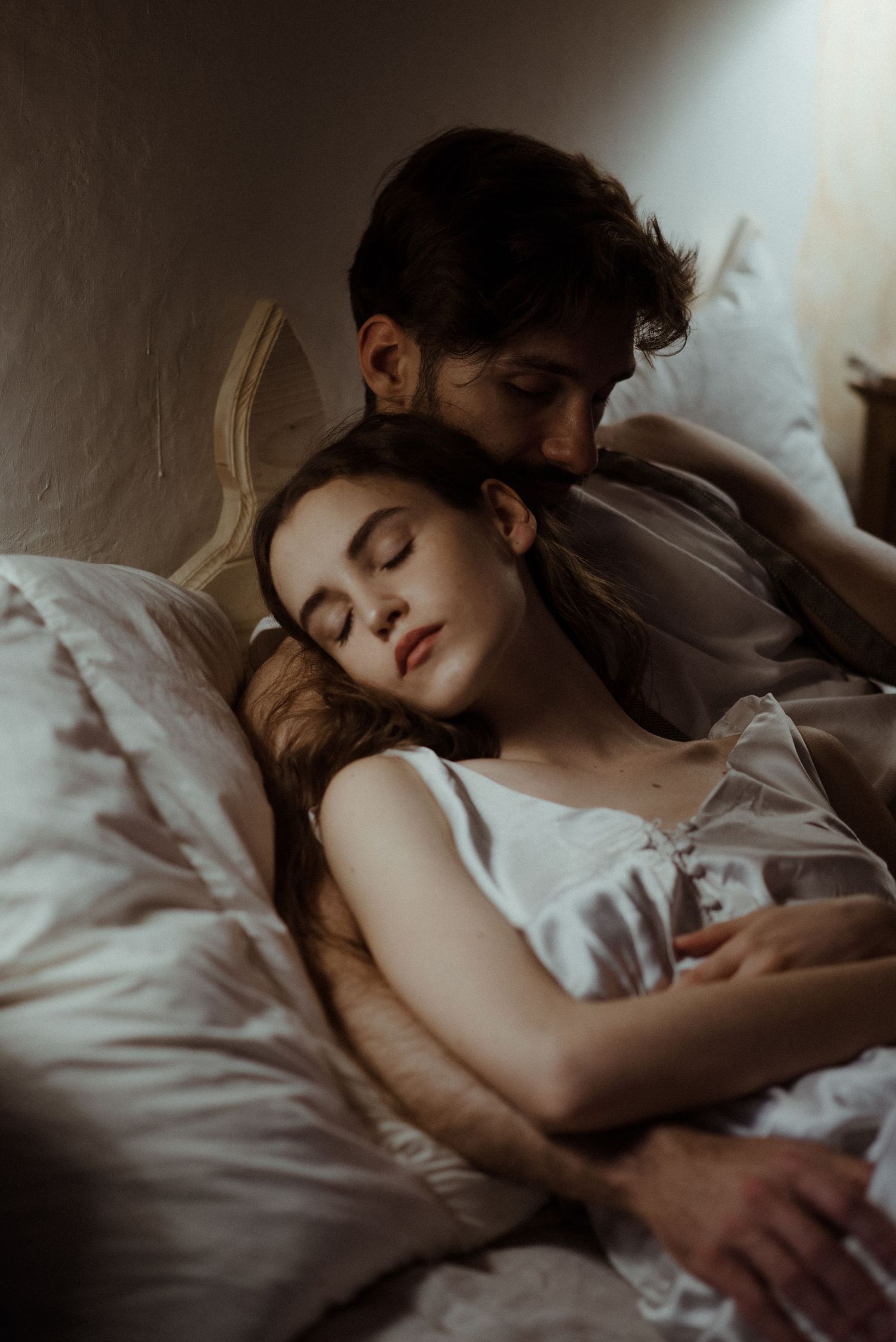 A couple shares an intimate moment while sleeping together in bed, bathed in soft natural light.