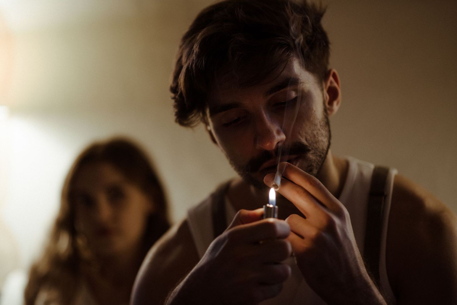 Person lighting a cigarette in dim lighting with silhouette of another person visible in background.