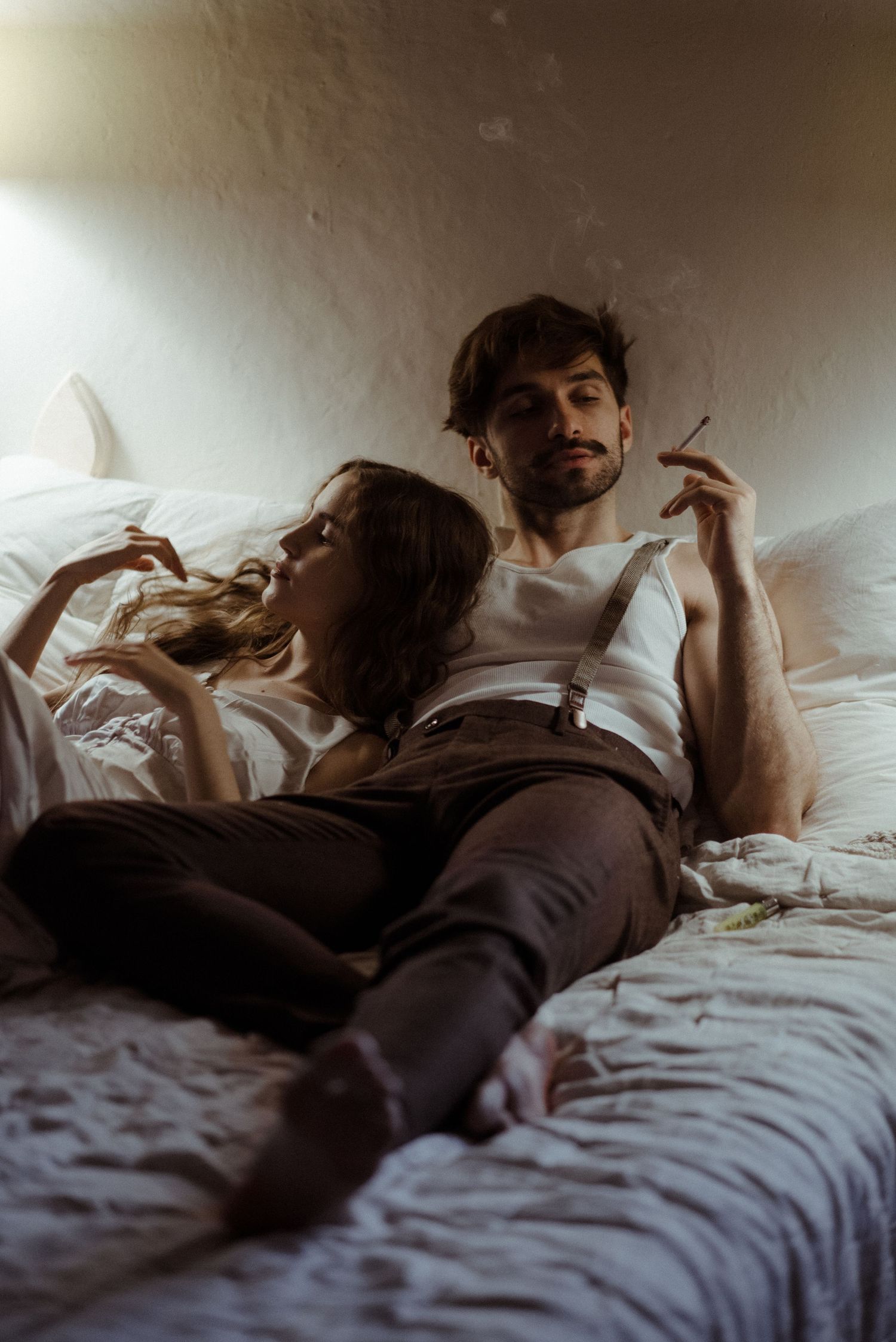 Couple relaxing together on white bedding in moody lighting creating an intimate atmospheric scene.