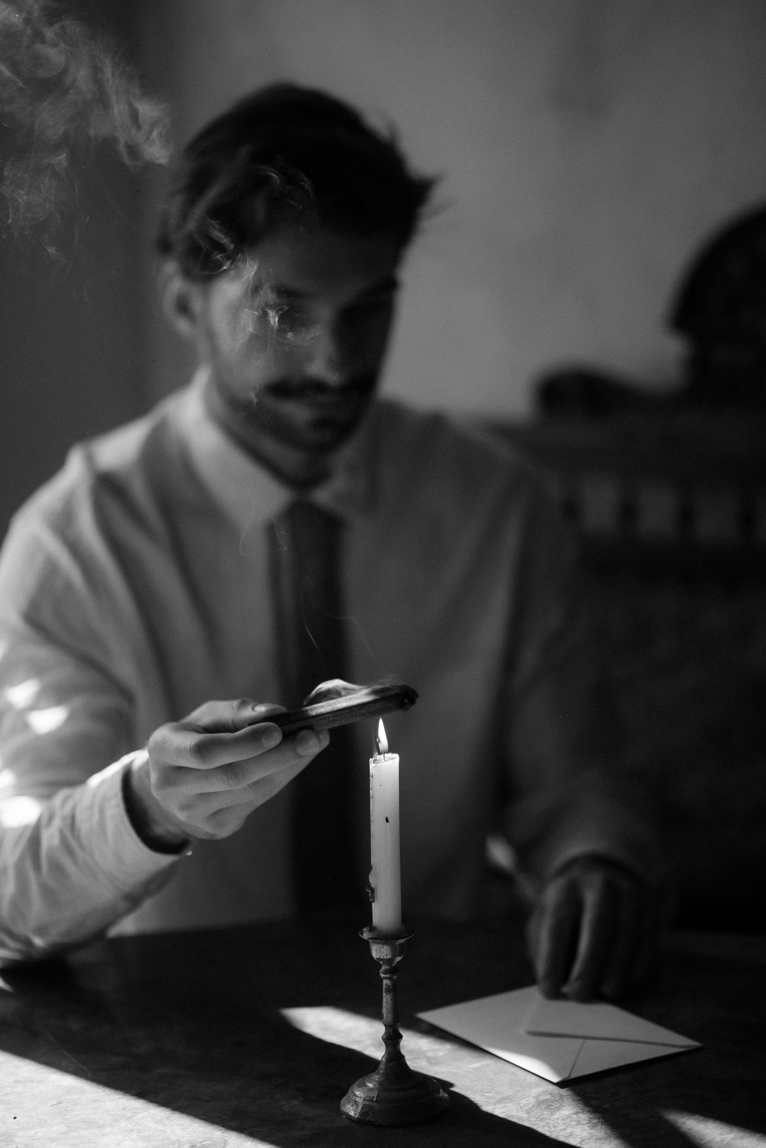 A black and white photo of someone lighting a cigarette from a candle while wearing a dress shirt and tie.