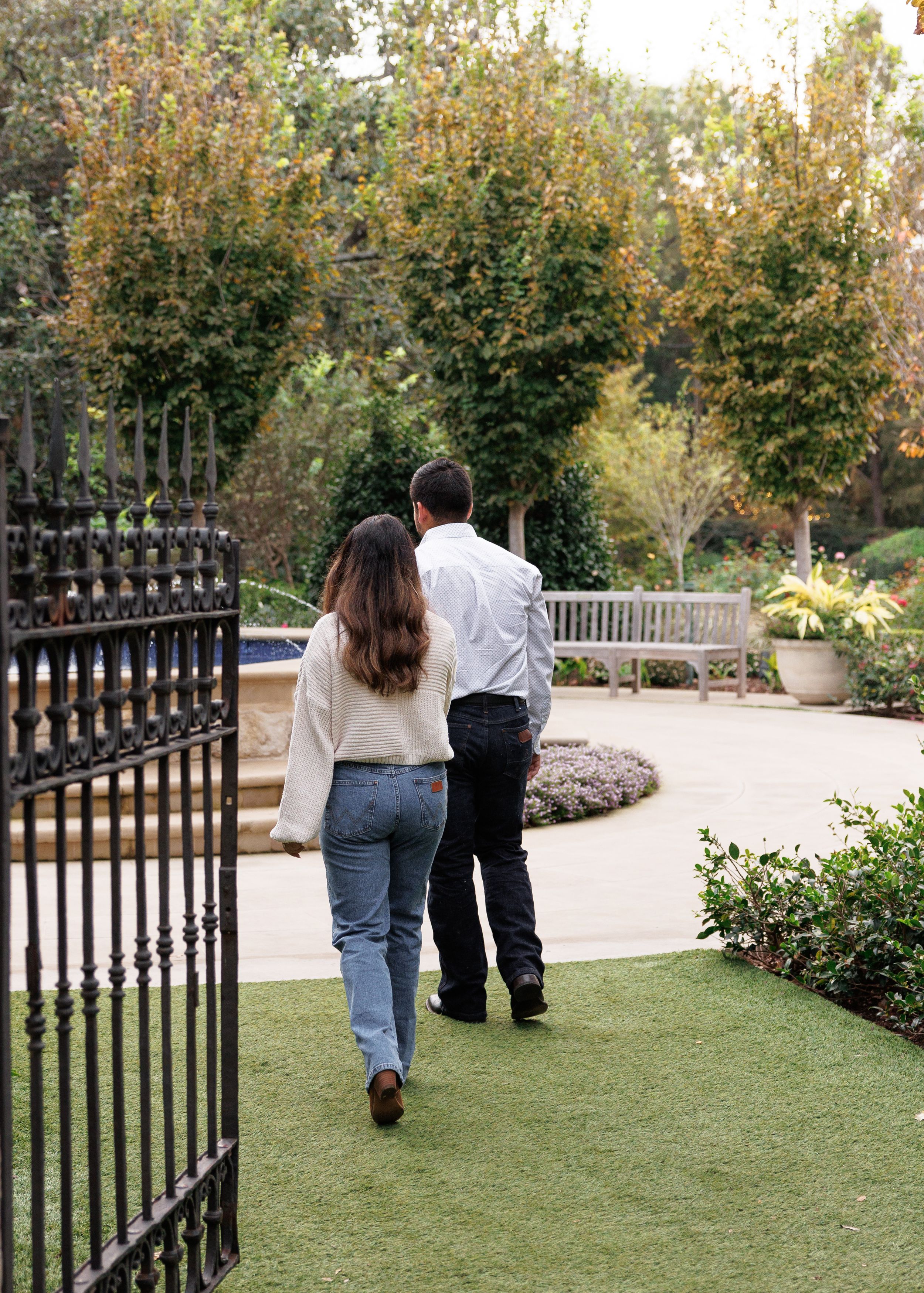 Two people walk along a garden path with wrought iron fence and lush greenery in the background.