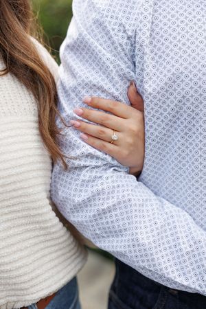 Close up of an engagement ring shown on a hand resting against a light blue patterned dress shirt and cream sweater.