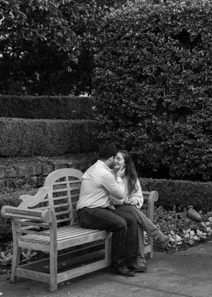A couple shares a tender moment while sitting together on a wooden garden bench surrounded by hedges.