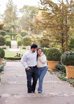 A couple walks together on a garden path surrounded by manicured hedges and autumn foliage at a formal estate.