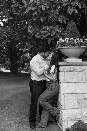 A romantic black and white photo of a couple sharing an intimate moment beside a stone pillar in a garden setting.