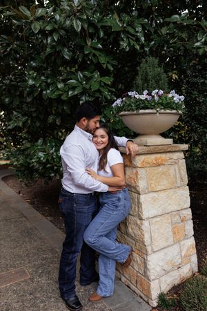 A couple shares a tender moment next to a stone pillar with a decorative planter in a garden setting.