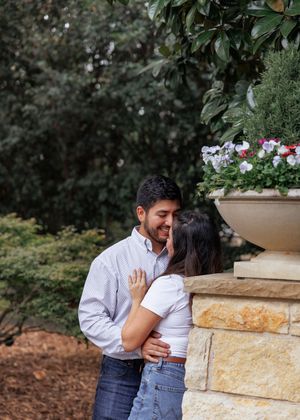 A couple embraces and shares a romantic moment near a stone pillar with flowers in an outdoor garden setting.