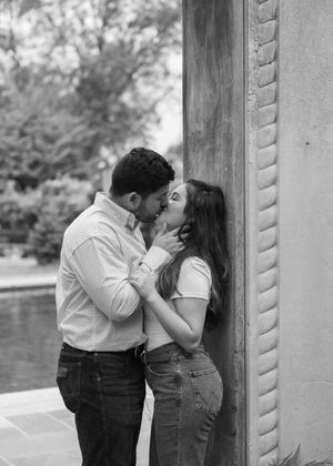 A romantic couple shares an intimate kiss against a stone pillar in a black and white photograph.