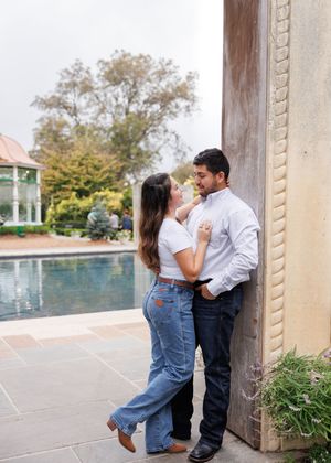 A couple embraces against an ornate pillar with a pool and garden in the background.