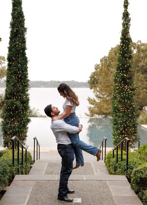 A romantic couple embraces on stone steps flanked by tall cypress trees decorated with Christmas lights by a lake.