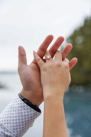 Hands from two people holding each other tenderly against blurred waterfront background, one wearing an engagement ring.