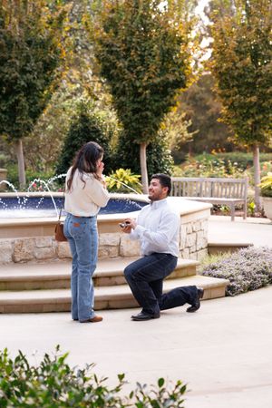 A romantic marriage proposal scene takes place in a garden setting with a fountain and autumn trees in the background.