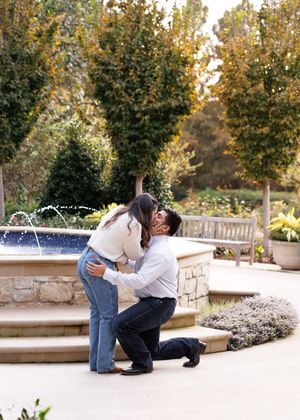 Beautiful proposal moment captured at a decorative stone fountain in a garden with autumn trees in the background.