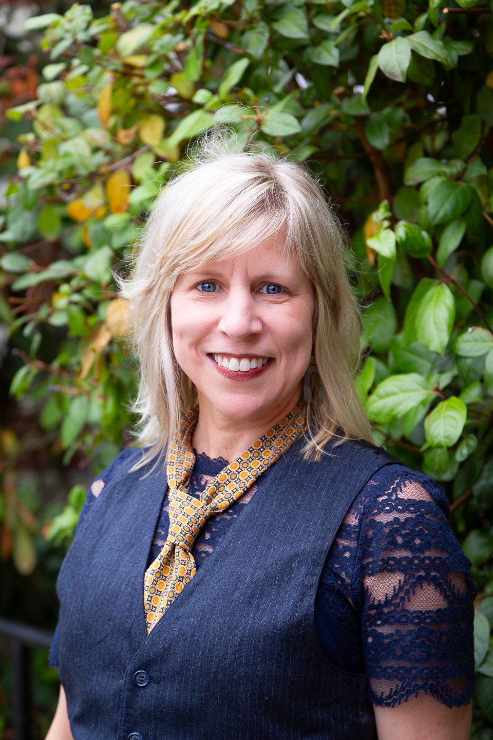 Professional outdoor portrait in front of green foliage featuring a navy vest and lace blouse with yellow scarf accessory.