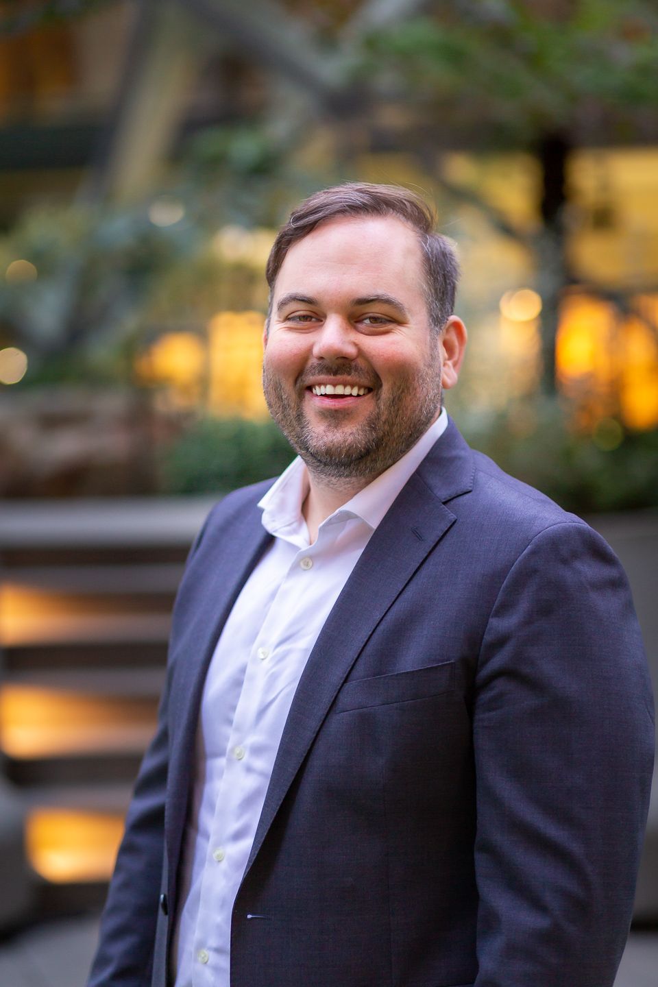 Professional headshot of someone in a navy suit and white shirt smiling outdoors with blurred background lighting.