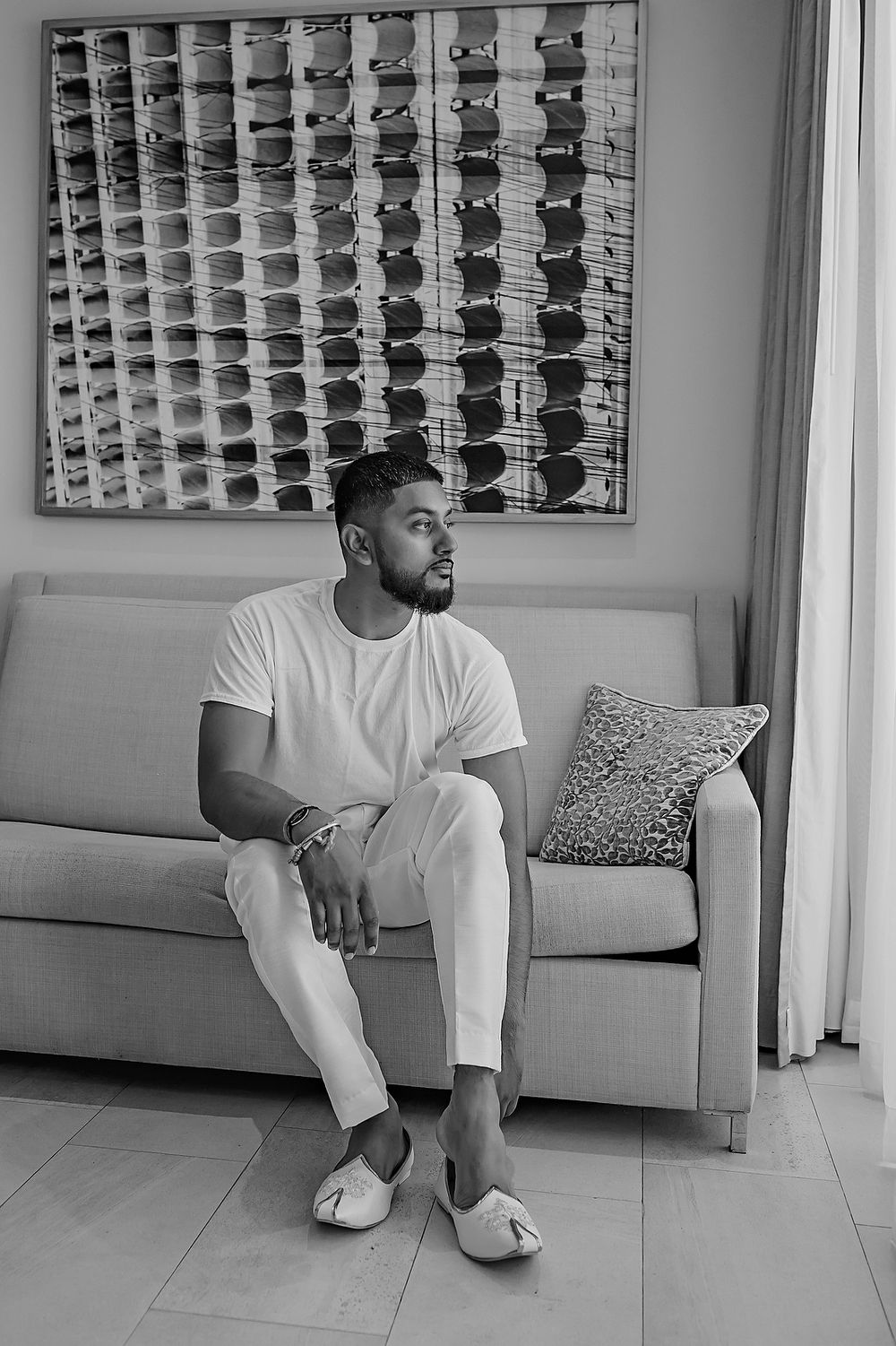 Black and white portrait of person in white clothing sitting on grey couch with decorative wall art in modern interior setting.
