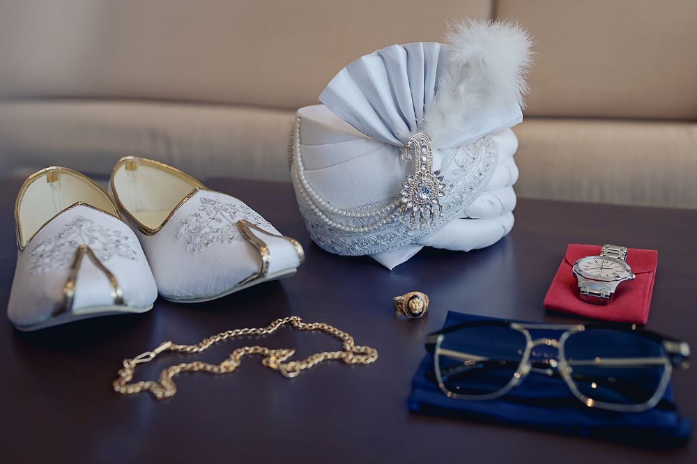 Wedding accessories including white turban, shoes, gold necklace, glasses and ring box displayed on dark surface.