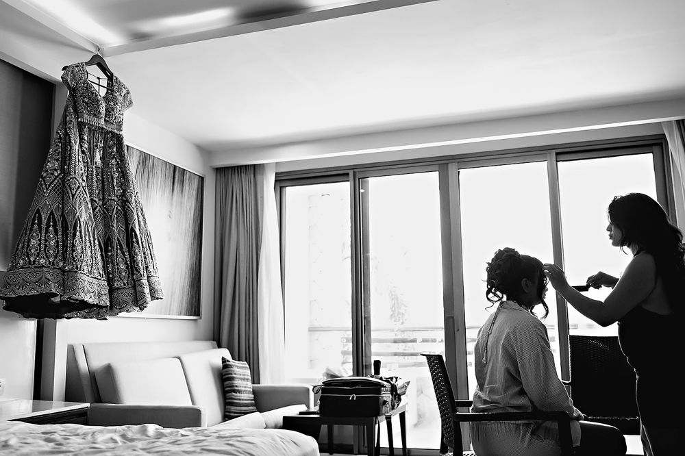 Black and white photo showing an elegant dress hanging in a room while a hairstyling session takes place by a window.