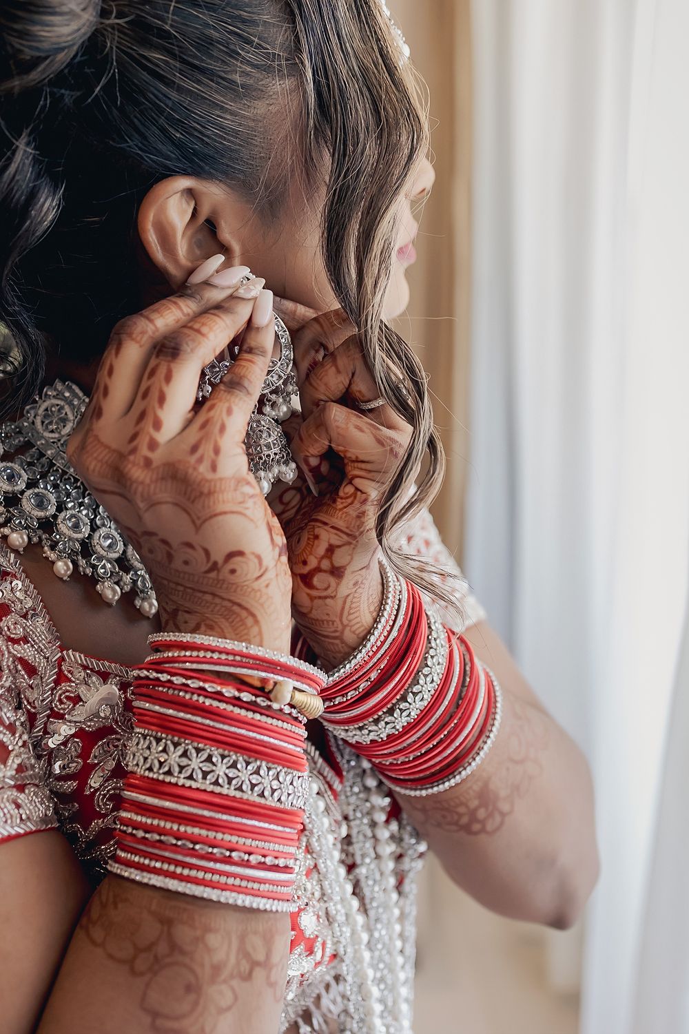 Traditional Indian bride wearing ornate jewelry and red and white bangles adjusts her earring in morning light.