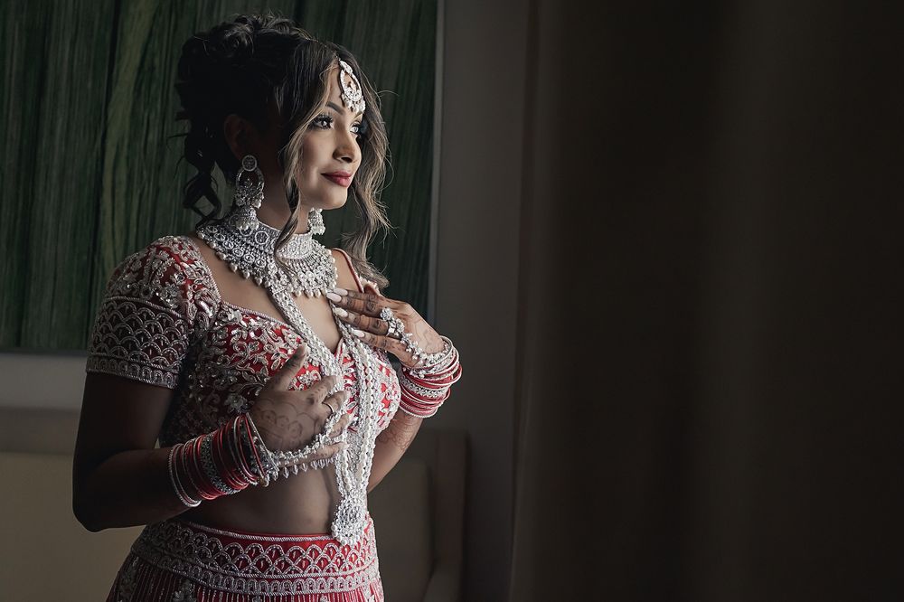 A bride in an ornate pink and white beaded wedding dress stands by a window in soft natural lighting.