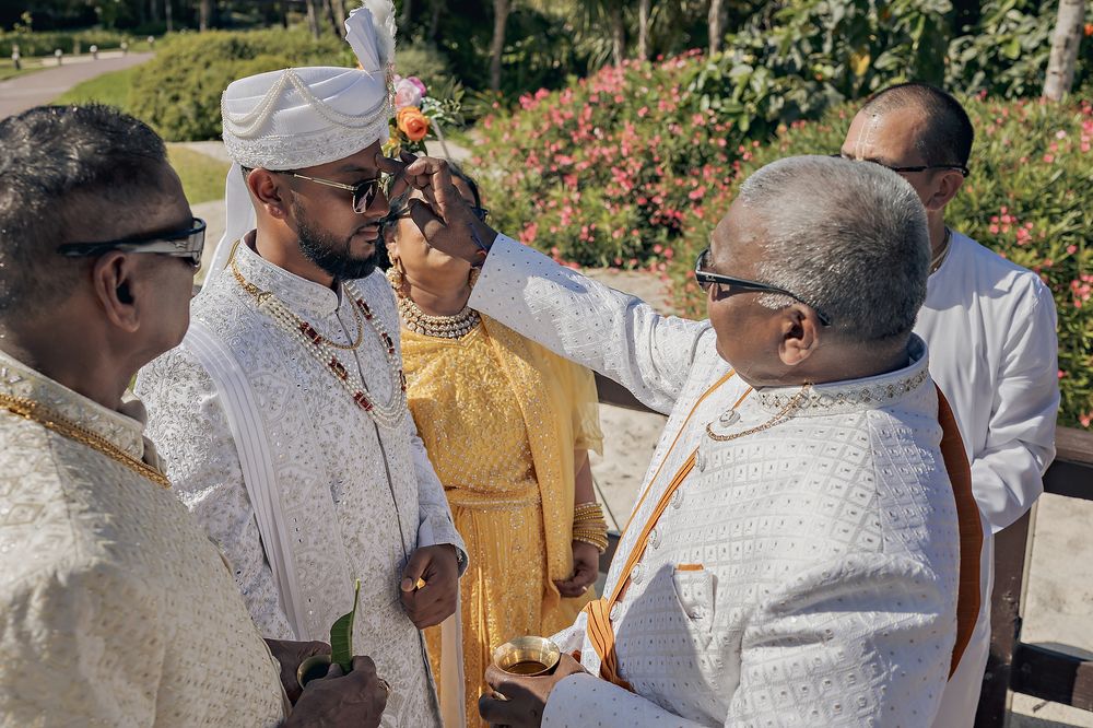 Two men in white formal attire perform a ceremonial blessing or ritual outdoors in a garden setting.