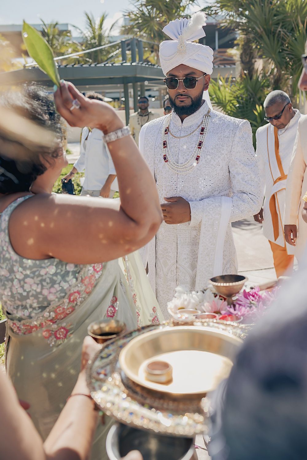 A person in traditional white wedding attire and turban participates in an outdoor ceremony with guests and decor.