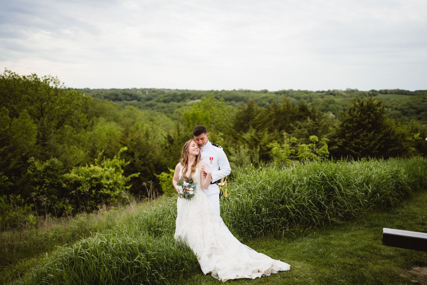 A couple embraces on a hilltop overlooking lush green countryside with the bride wearing a flowing white gown.