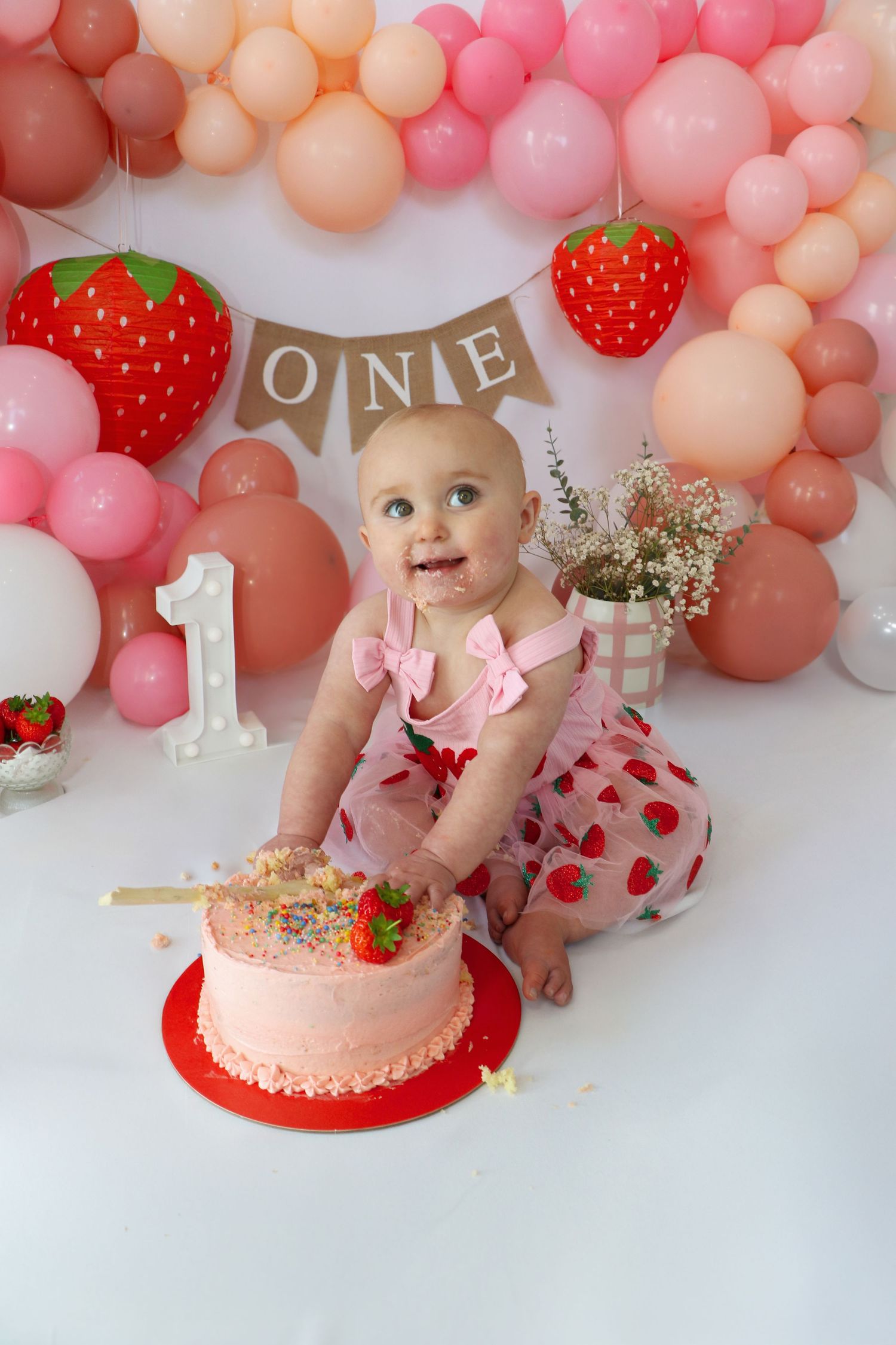 Baby in white dress touches birthday cake against white and grey balloon backdrop. Coventry Cake Smash Photographer. First Birthday