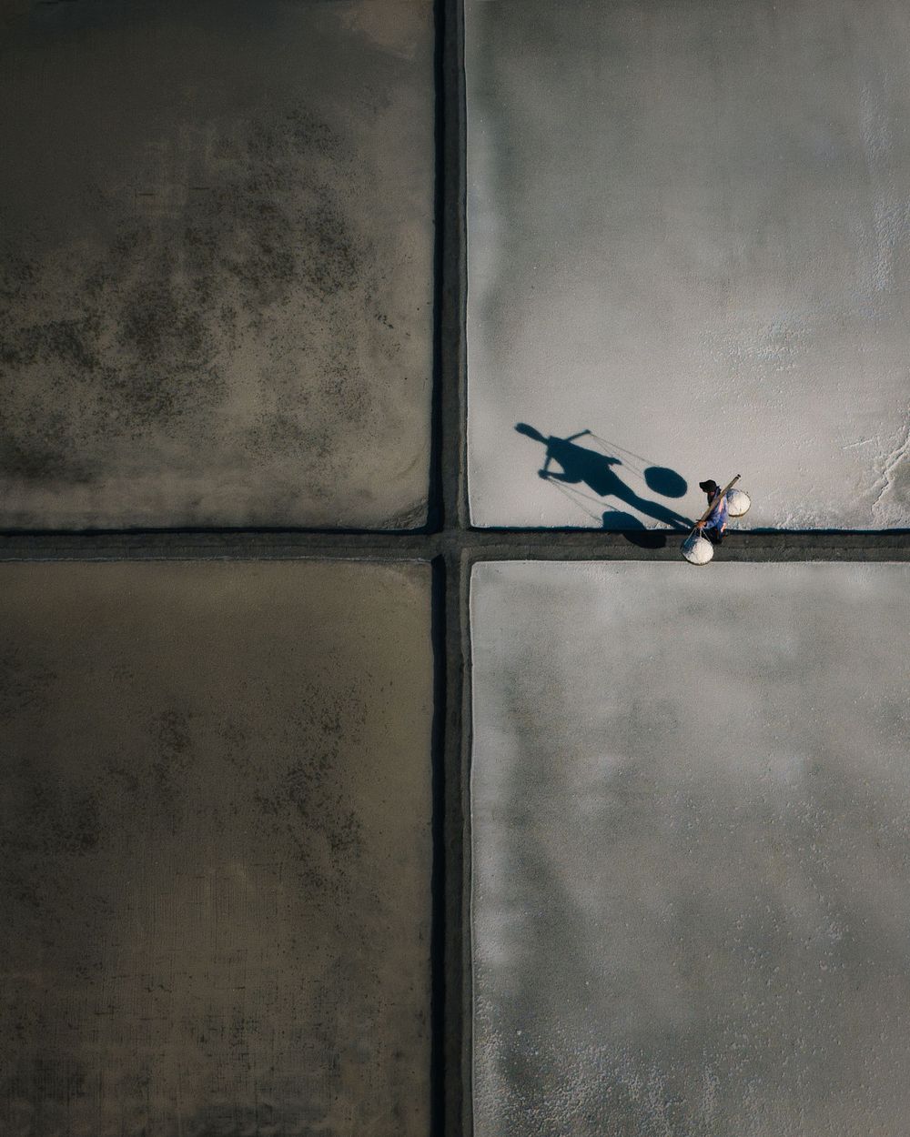 Aerial view of a person casting a long shadow on four concrete squares, creating a minimalist geometric composition.