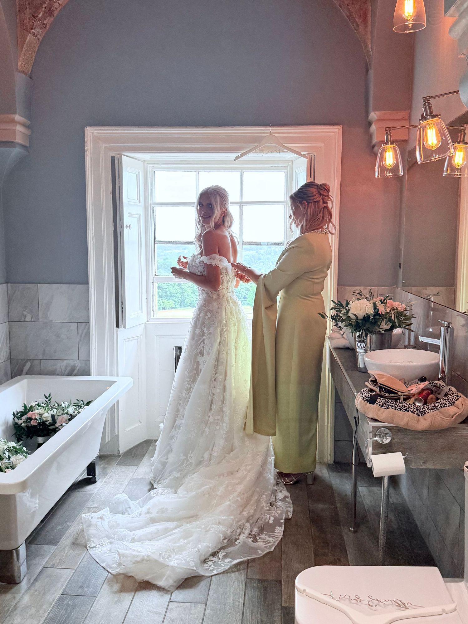 Mother of the bride in an elegant like green dress fastening her daughter’s ivory lace dress at the back.
