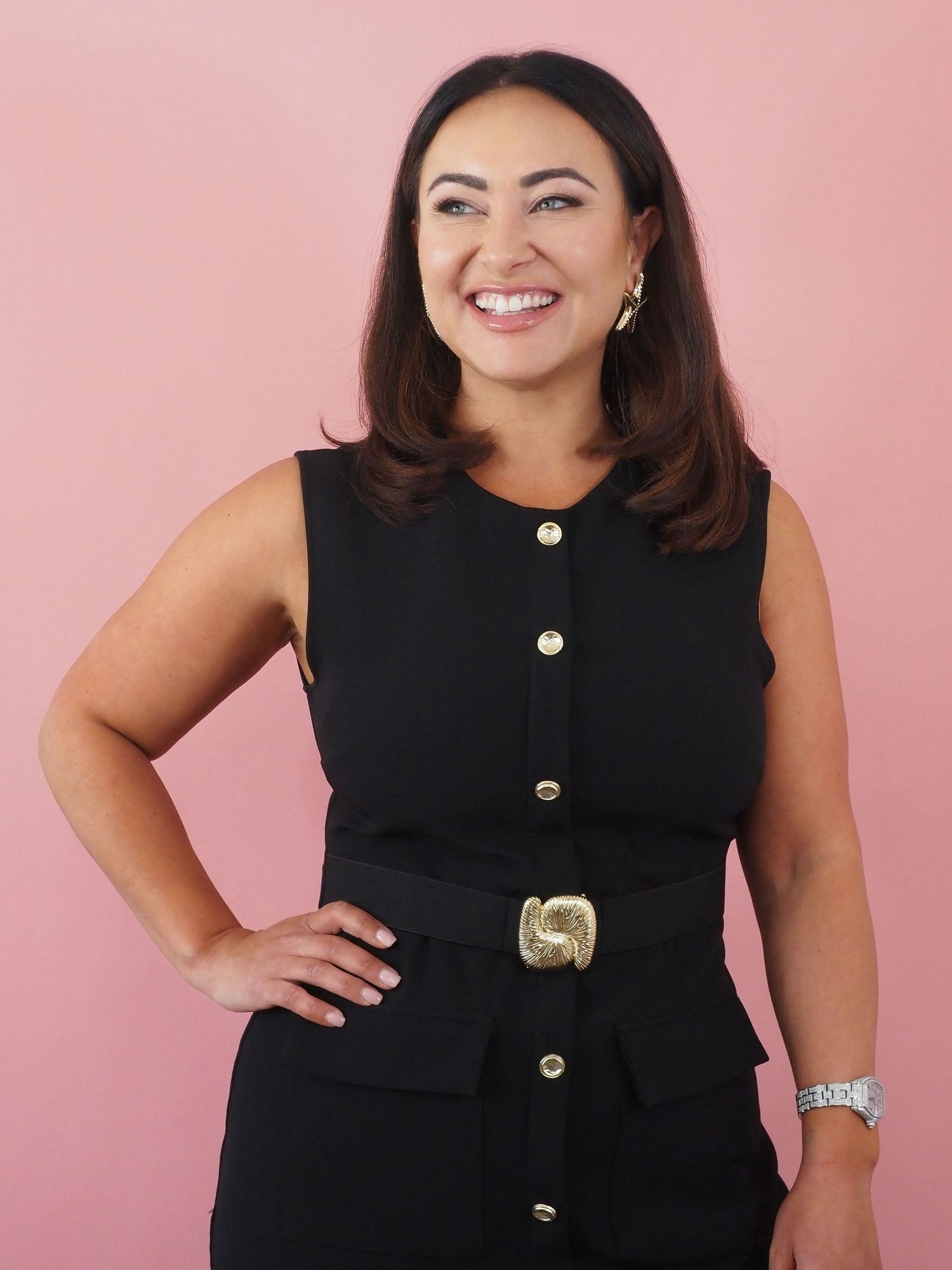 Joyful woman in a black sleeveless dress with gold buttons and belt buckle poses against a pink background.
