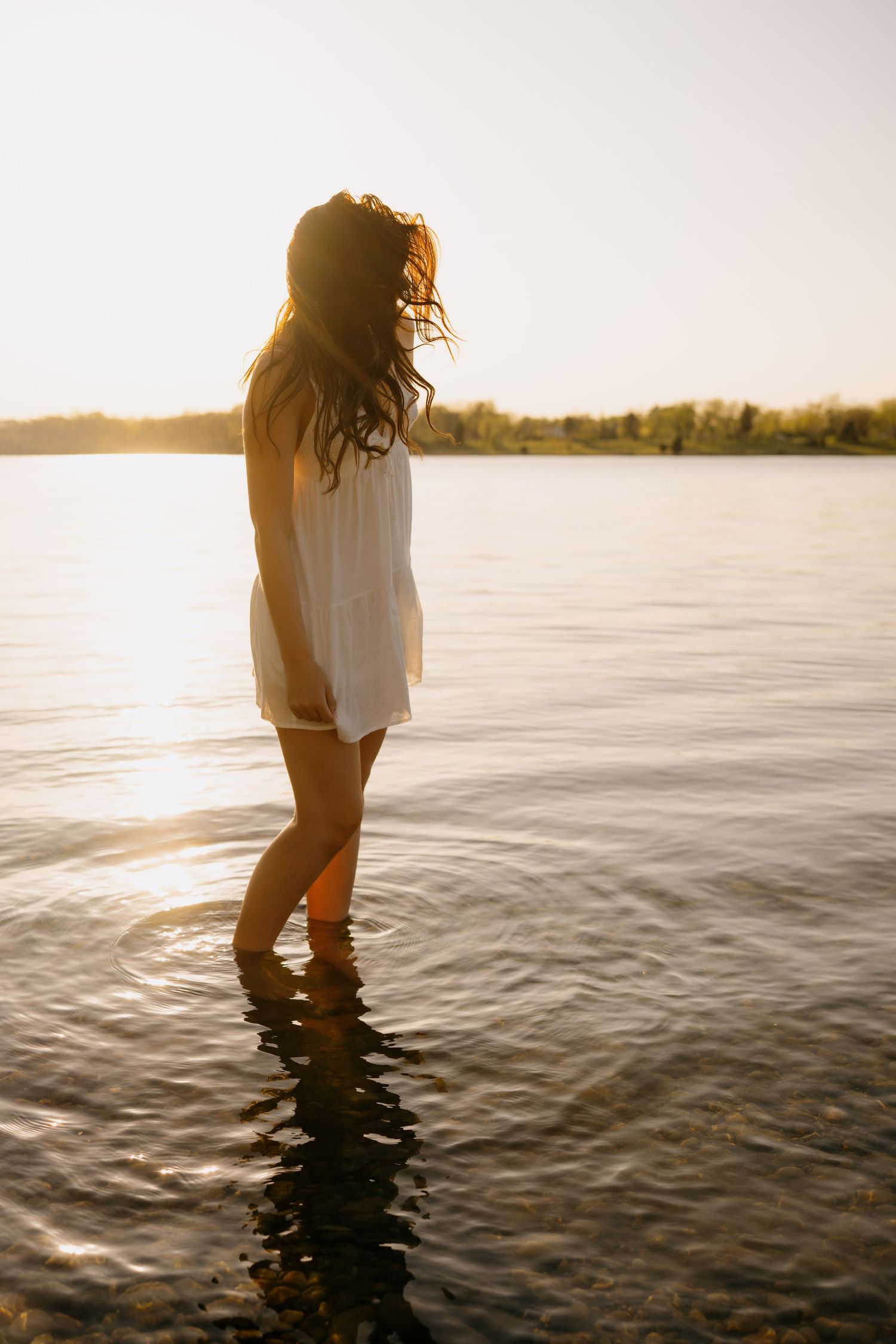 Silhouetted figure in white dress stands in calm lake water during golden sunset with trees in background.