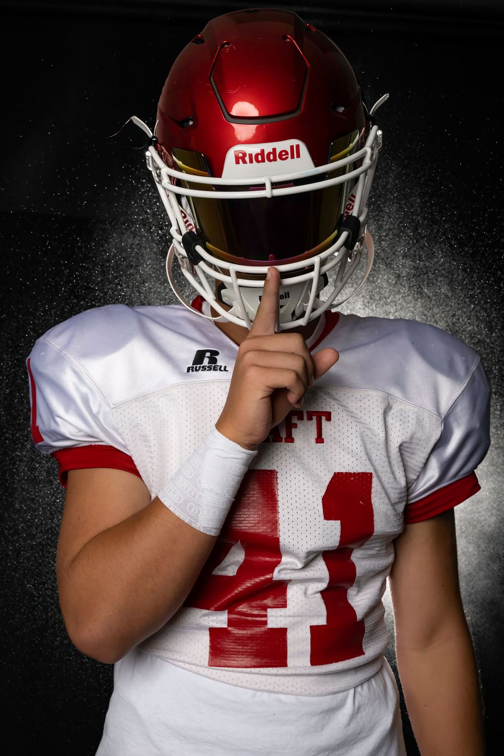 Football player in white jersey and red helmet holds finger to facemask in hushing gesture against dark background.