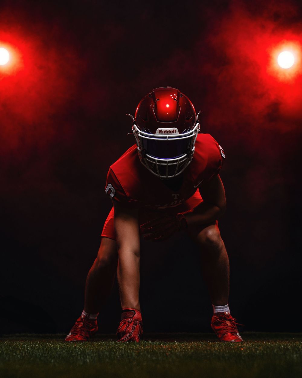 Football player in red jersey crouches in ready position on field at night with dramatic red lighting overhead.