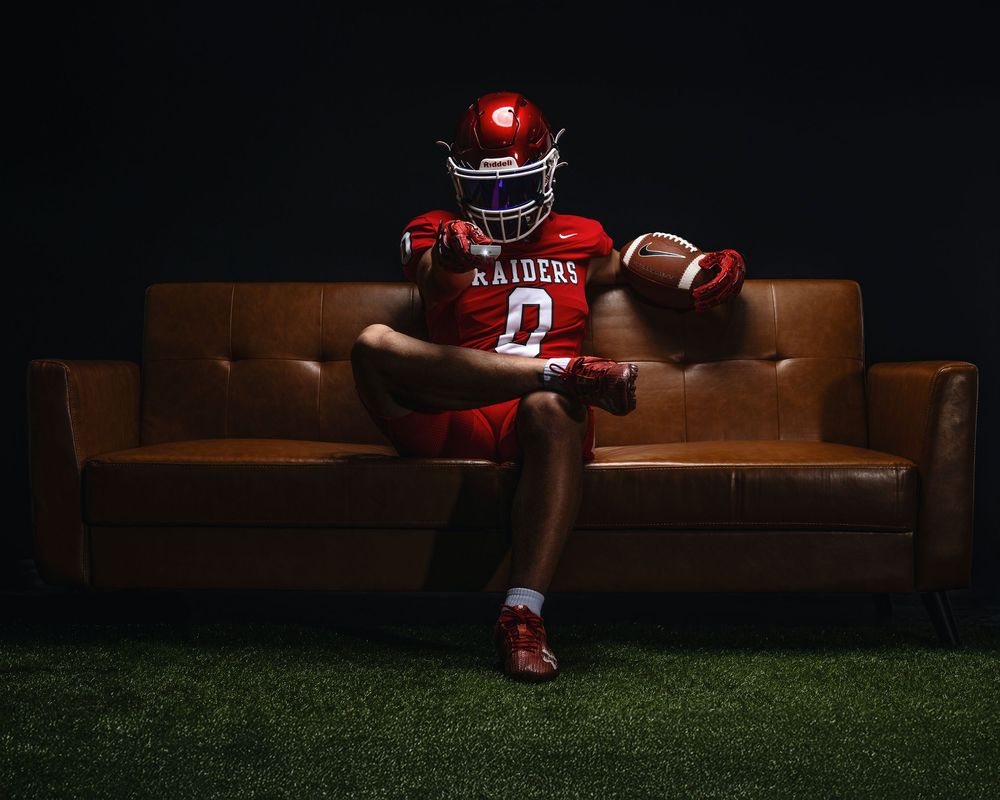 Football player in red uniform sits confidently on brown leather couch against dark background with artificial turf below.